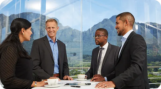 Four business professionals standing around a table, smiling and talking in a modern office with mountain views in the background.